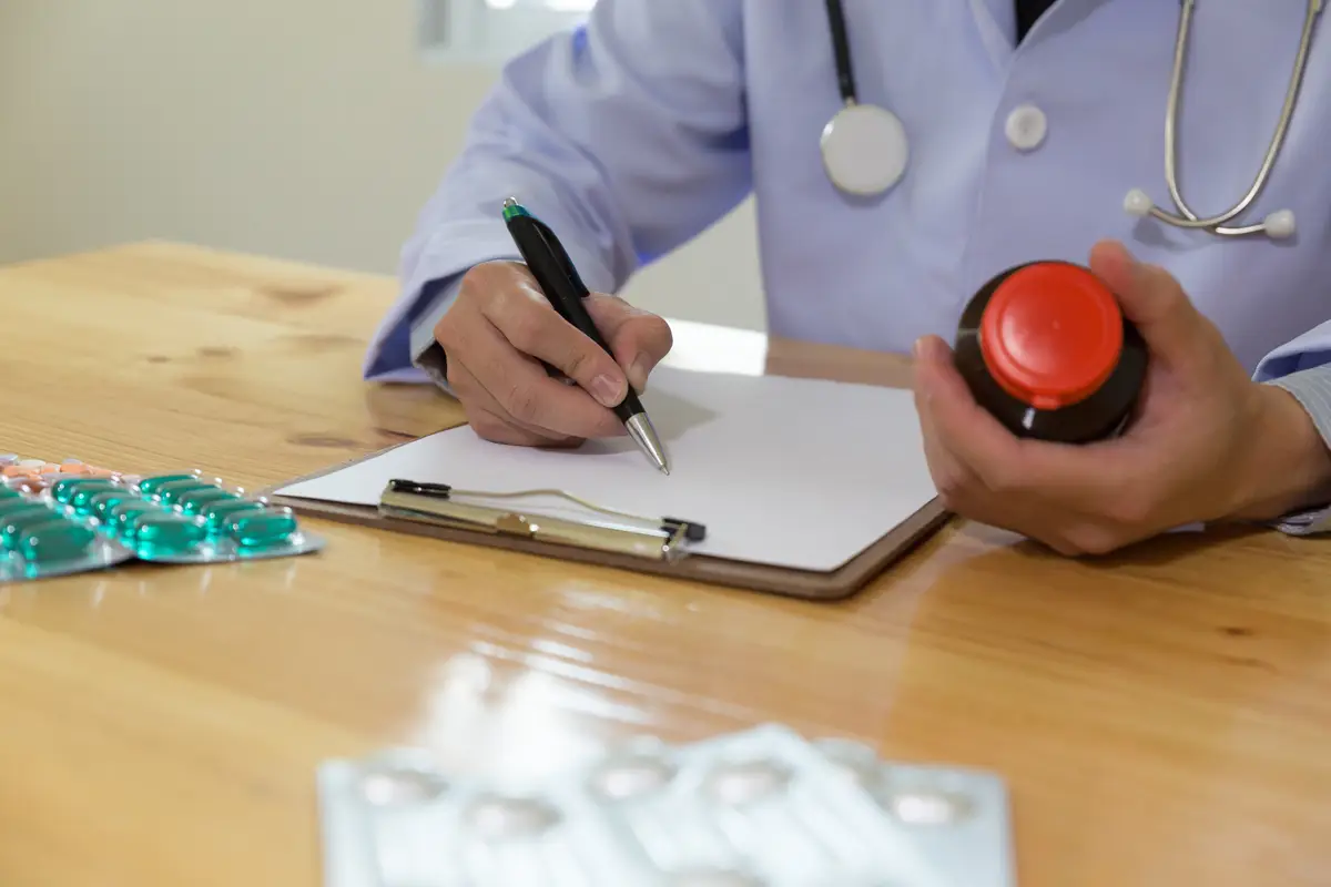 Midsection of doctor writing prescription while holding bottle on table