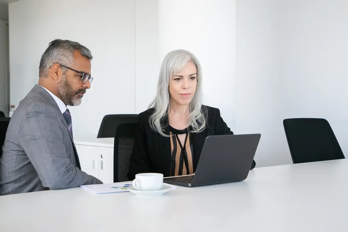 Friendly colleagues using laptop for video call, sitting at table with cup of coffee, looking at display and talking. Online communication concept