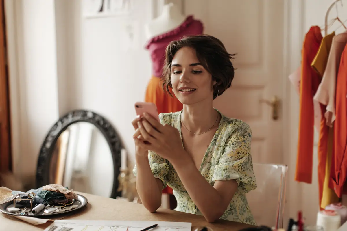 Cheerful tanned brunette shorthaired woman in floral vneck dress chatting on phone sits at desk and poses in cozy fashion designer office
