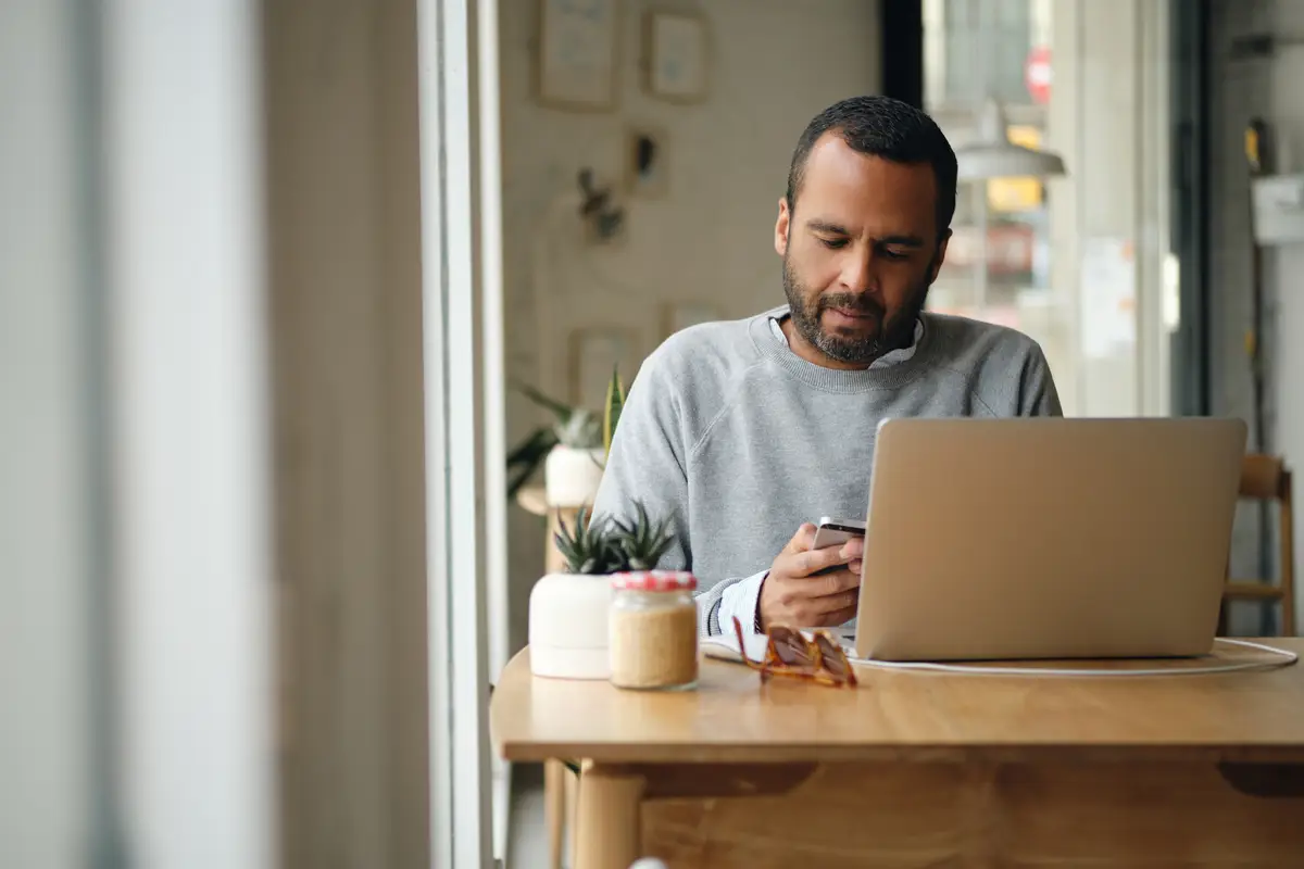 LLC nos EUA: Como Abrir, Vantagens e Erros a Evitar 3 Adult businessman in sweater intently using smartphone while working on laptop in city cafe