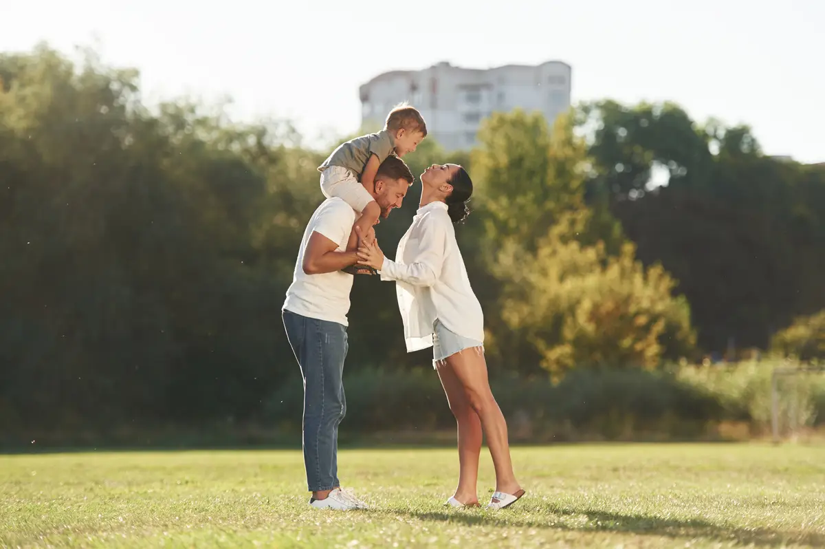 Kid is sitting on the shoulders Mother and father are with son outdoors on the field