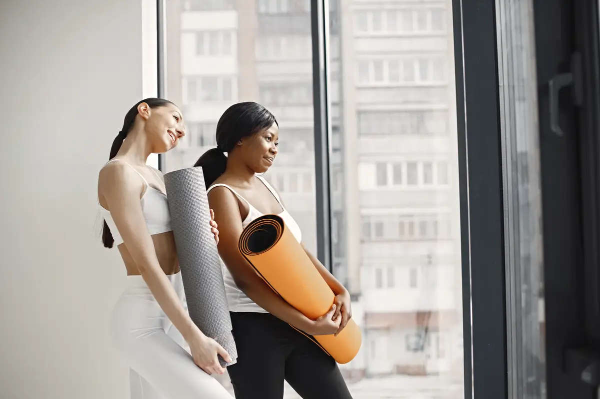 Black and caucasian women standing with rolling mats in studio with big windows