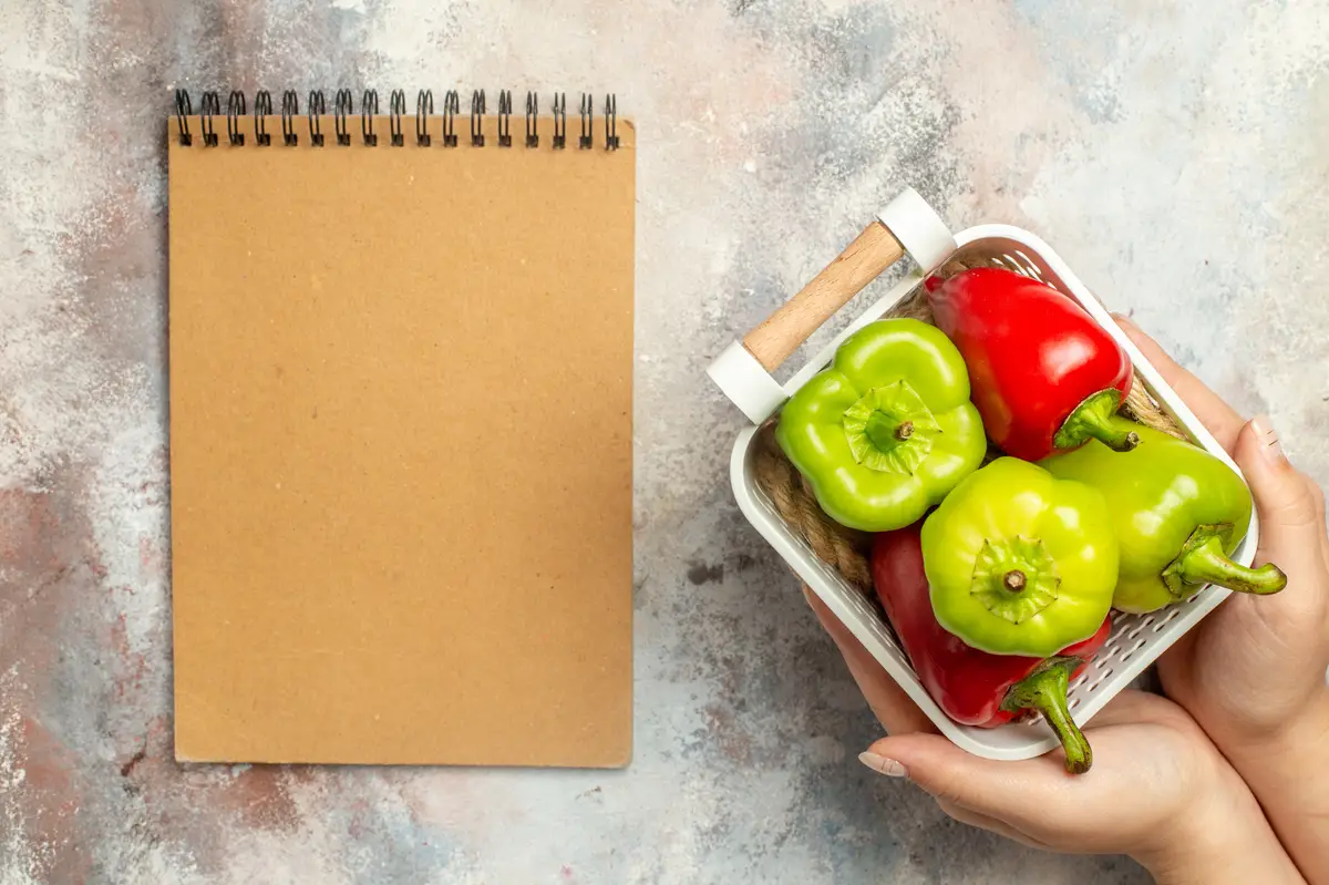 Top view green and red peppers in plastic basket in female hand a notepad on nude surface