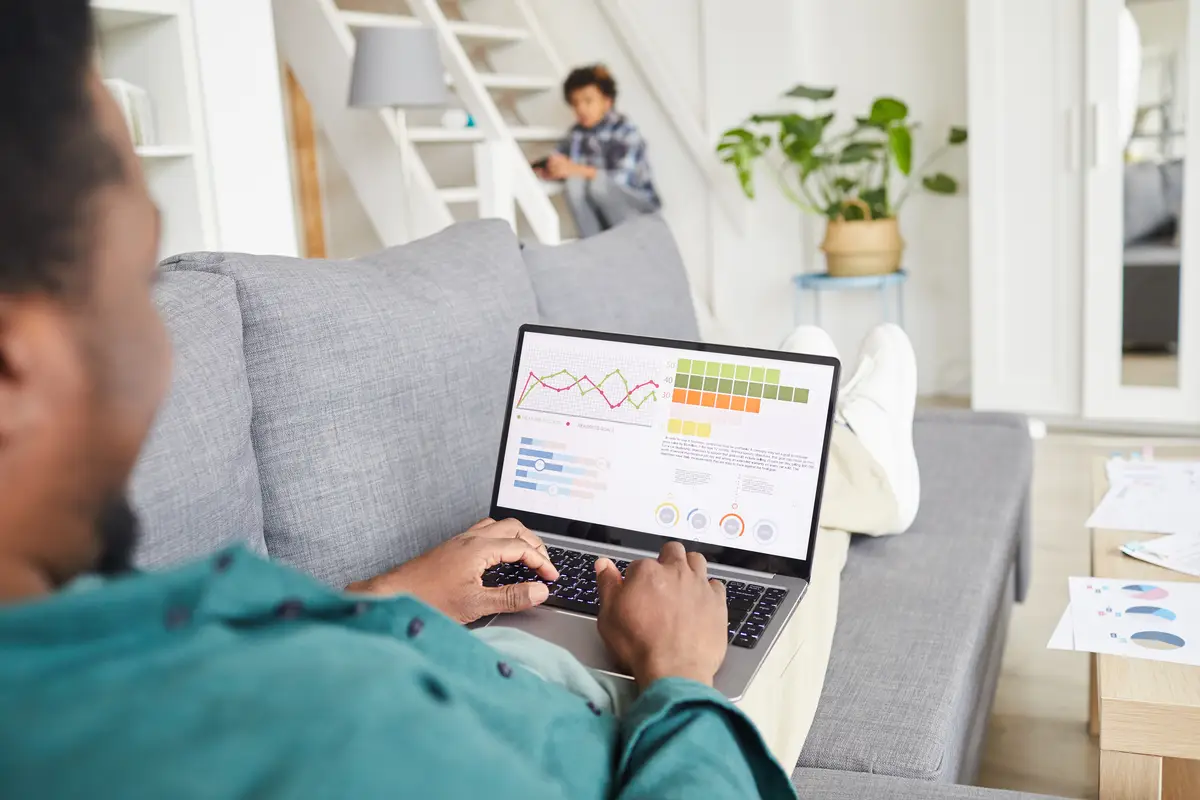 African young man lying on sofa with laptop on his knees and working online in the living room