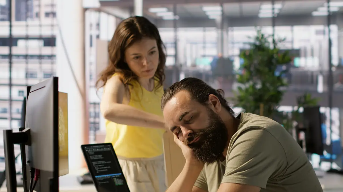 Tired worker caught asleep at desk in office by colleague laughing about it