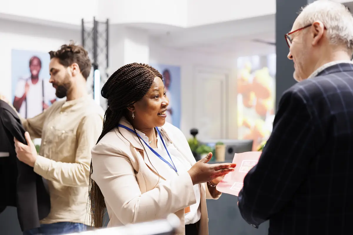 Friendly smiling African American woman shop sales assistant explaining senior man customer Black Friday shopping rules in clothing store. Retail worker holding sales flyer talking with shopper