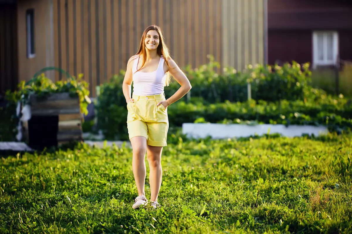 Smiling woman in his s in tank top and yellow shorts is walking in backyard of country house near