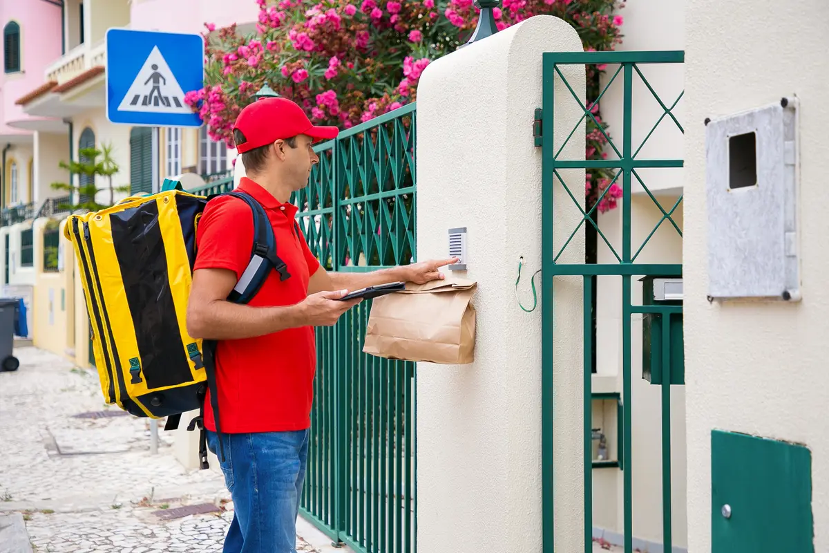 Endereço nos EUA: guia para brasileiros entenderem e utilizarem 2 Pensive deliveryman in red cap ringing in doorbell of recipient. Middle-aged courier with yellow thermal backpack delivering express order and standing on street. Delivery service and post concept