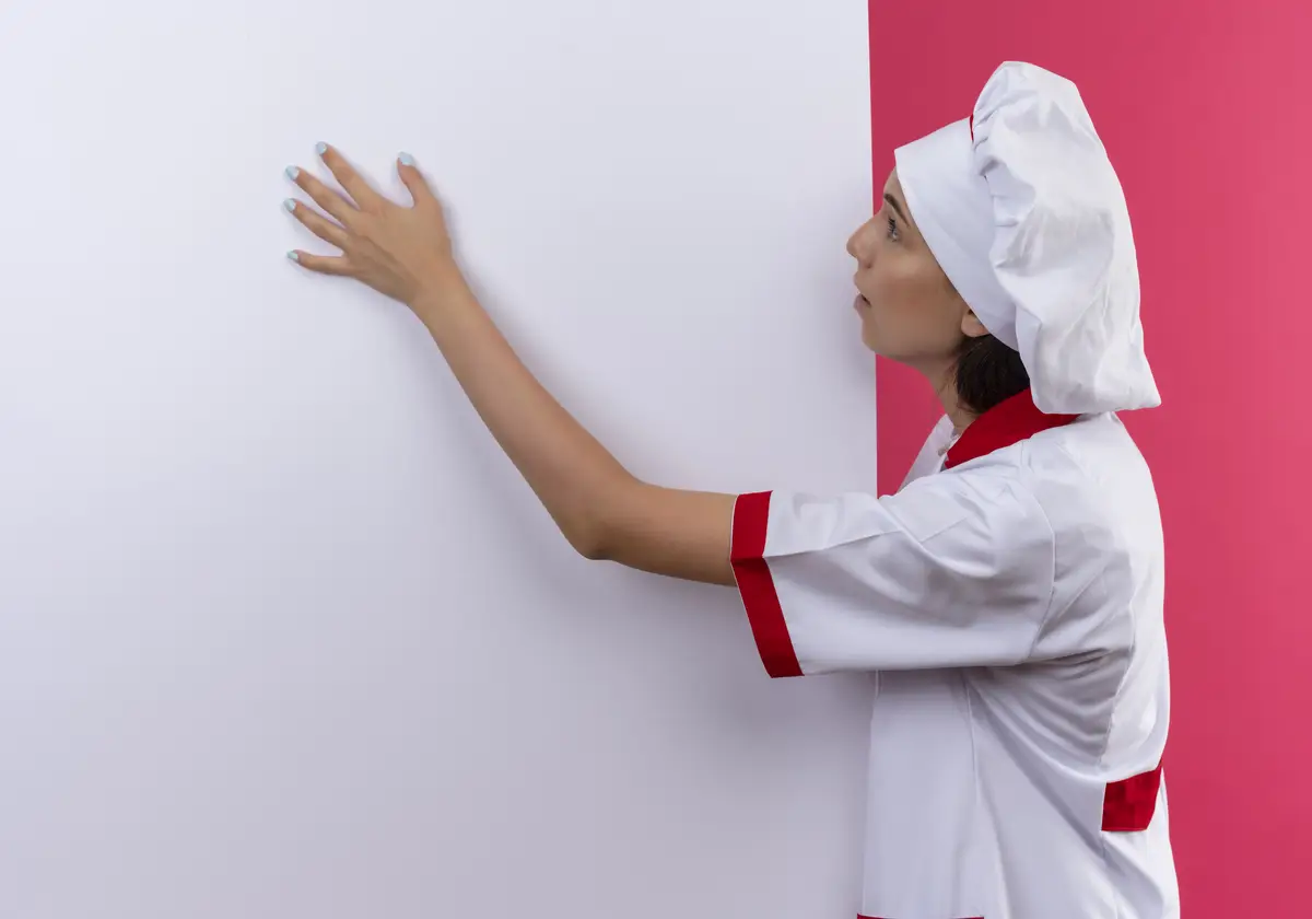 Young surprised caucasian cook girl in chef uniform looks and puts hand on white wall on pink  with copy space