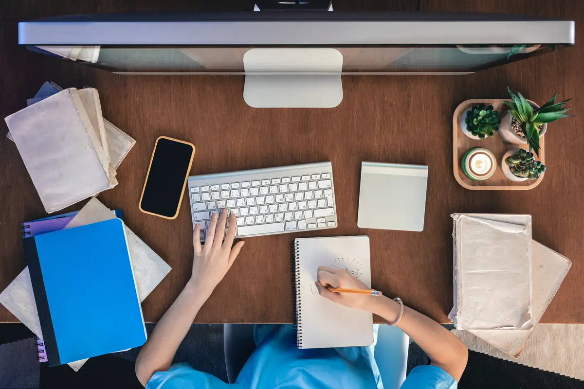 A girl sits in front of a computer with notepads and books top view