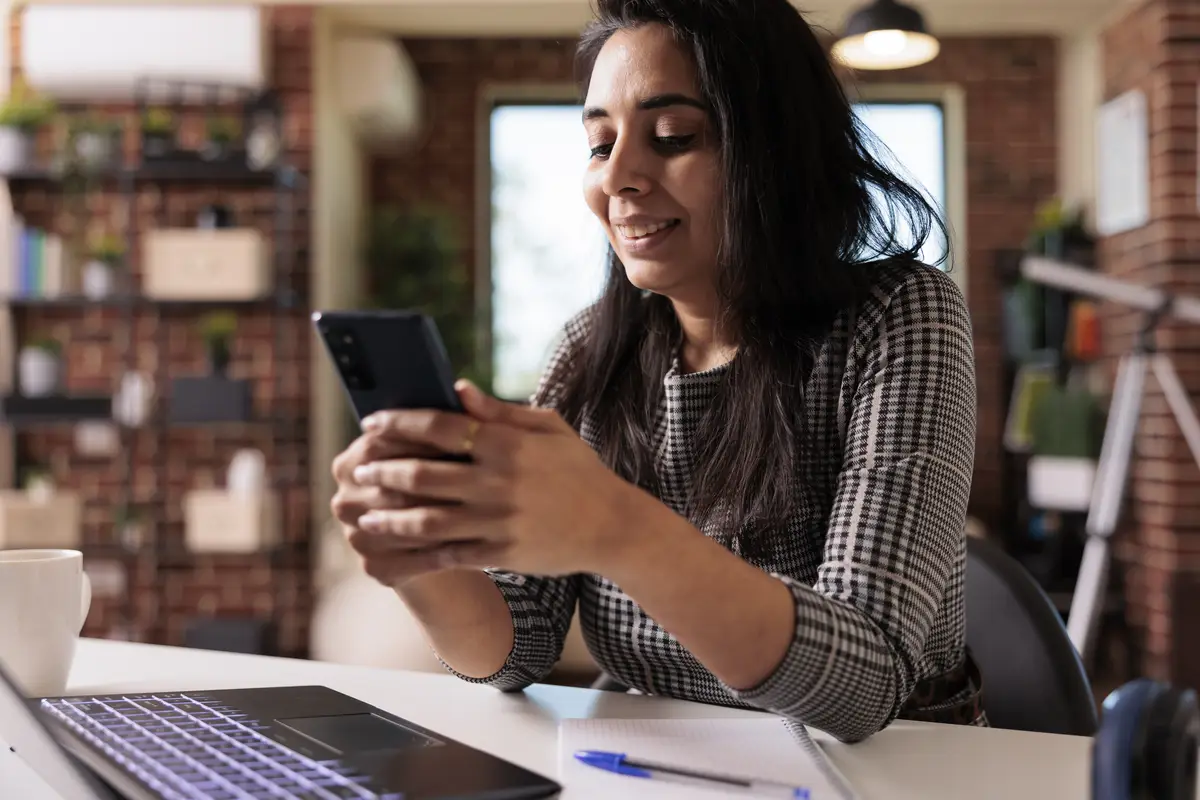 Female freelancer working on smartphone app and using laptop for remote job at home desk. Browsing internet and social media on mobile phone to text messages, online research break.