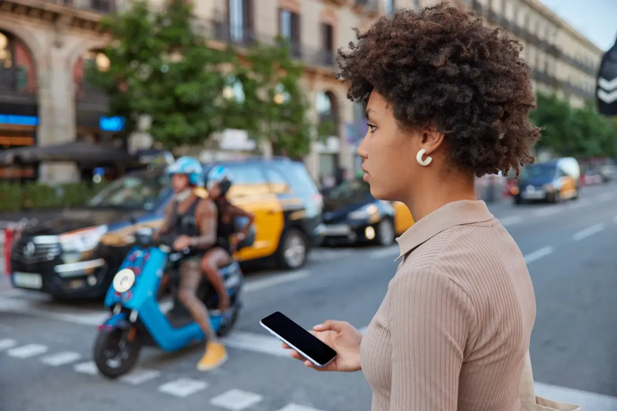 Curly haired female pedestrian crosses busy road holds modern smartphone with blank mockup screen poses outdoors