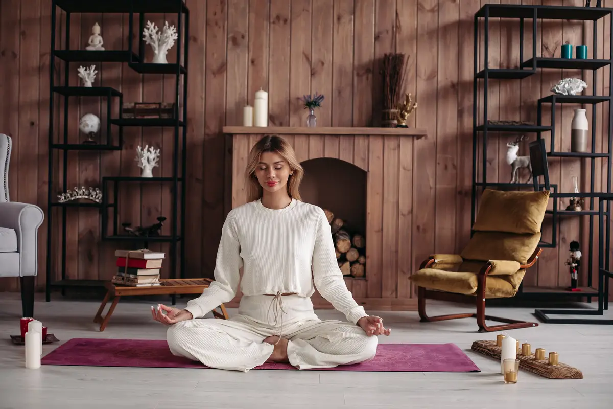 Smiling young girl practicing yoga sitting in lotus pose, meditating in cozy home interior. Female training for wellness. 