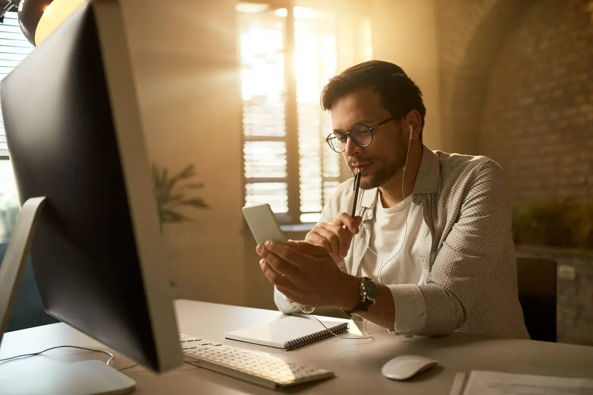 Pensive entrepreneur texting on mobile phone while working on a computer at his office desk