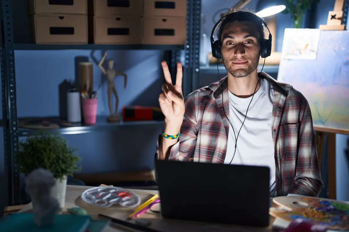 Young hispanic man sitting at art studio with laptop late at night showing and pointing up with fingers number two while smiling confident and happy