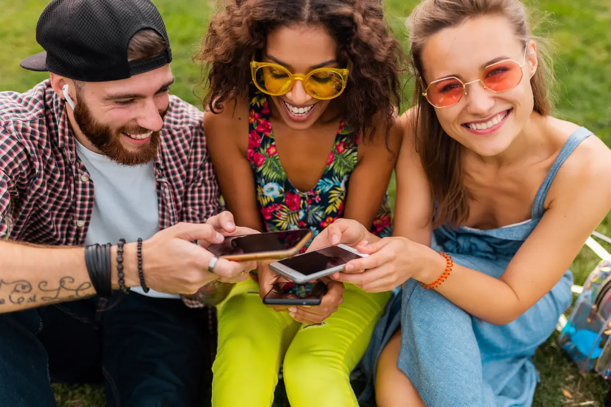 Happy young company of smiling friends sitting park using smartphones, man and women having fun together