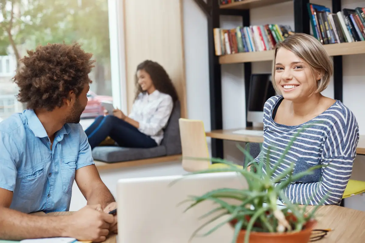 Group of three young good-looking multi-ethnic students sitting in university library. Dark-skinned guy looking back at his friend reading article. Light-haired girl with happy face