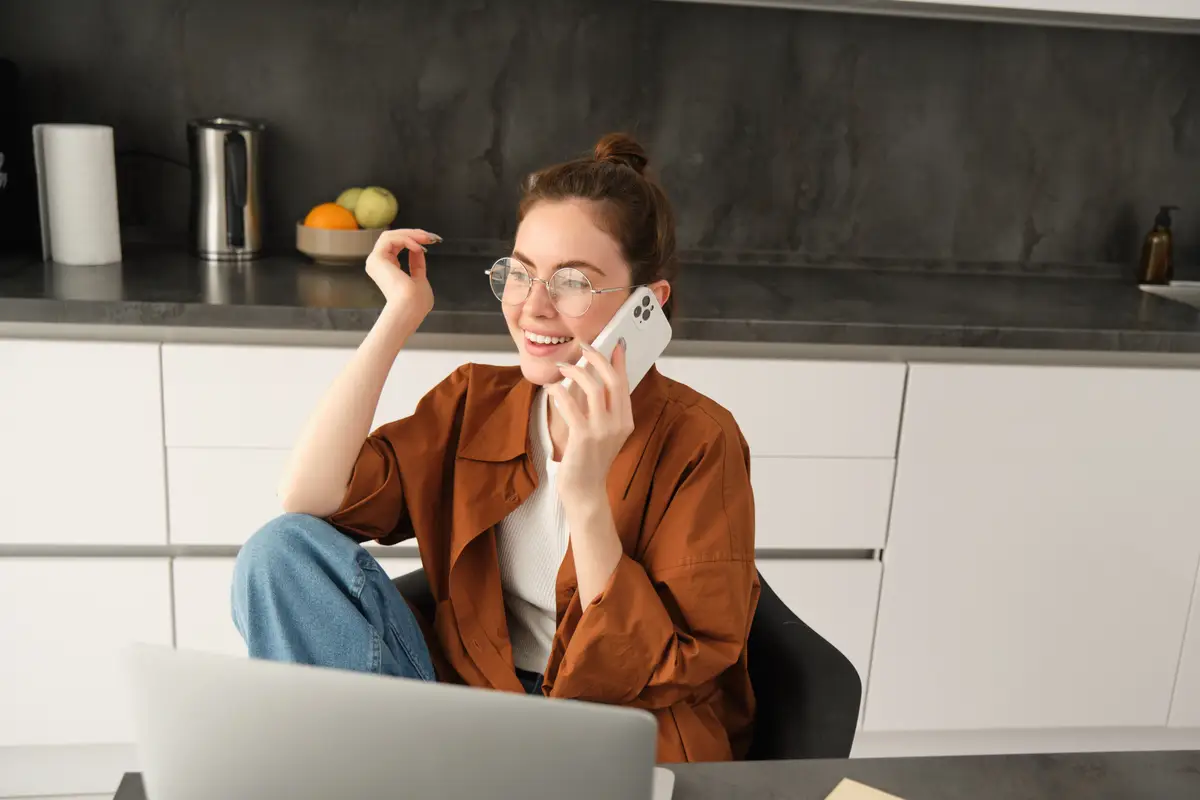 Excited young woman talking on mobile phone in front of laptop sitting in kitchen with happy face