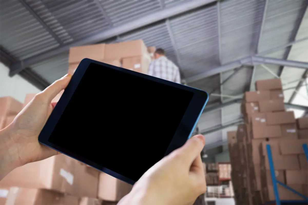Hands holding tablet against rear view of worker on ladder in warehouse