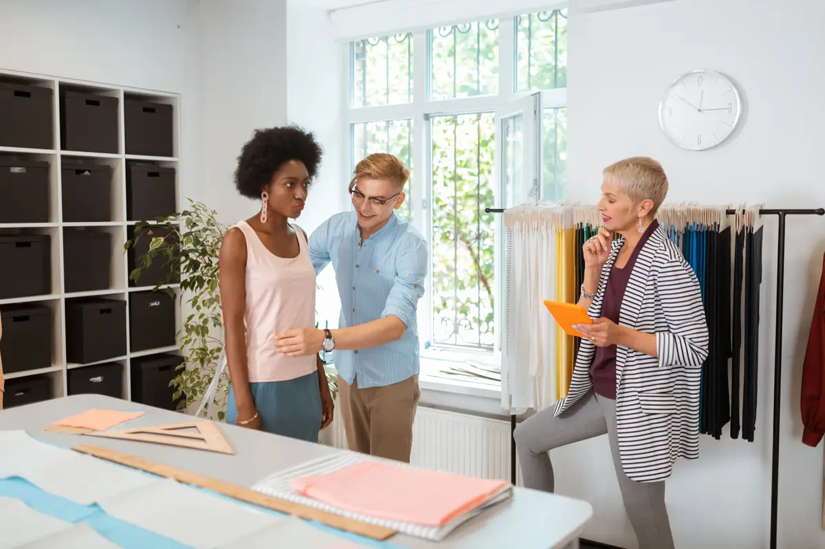 Joyful handsome tailor measuring a serious young woman looking at a smiling female fashion designer