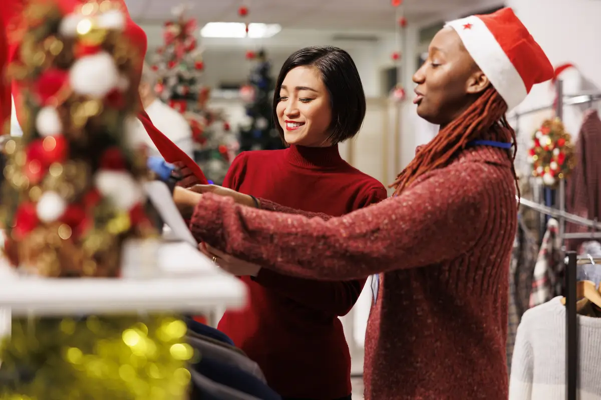 Store clerk helping asian client to find right measurement, recommending clothes and accessories for christmas dinner preparations at mall. Diverse women discussing about fabrics quality.