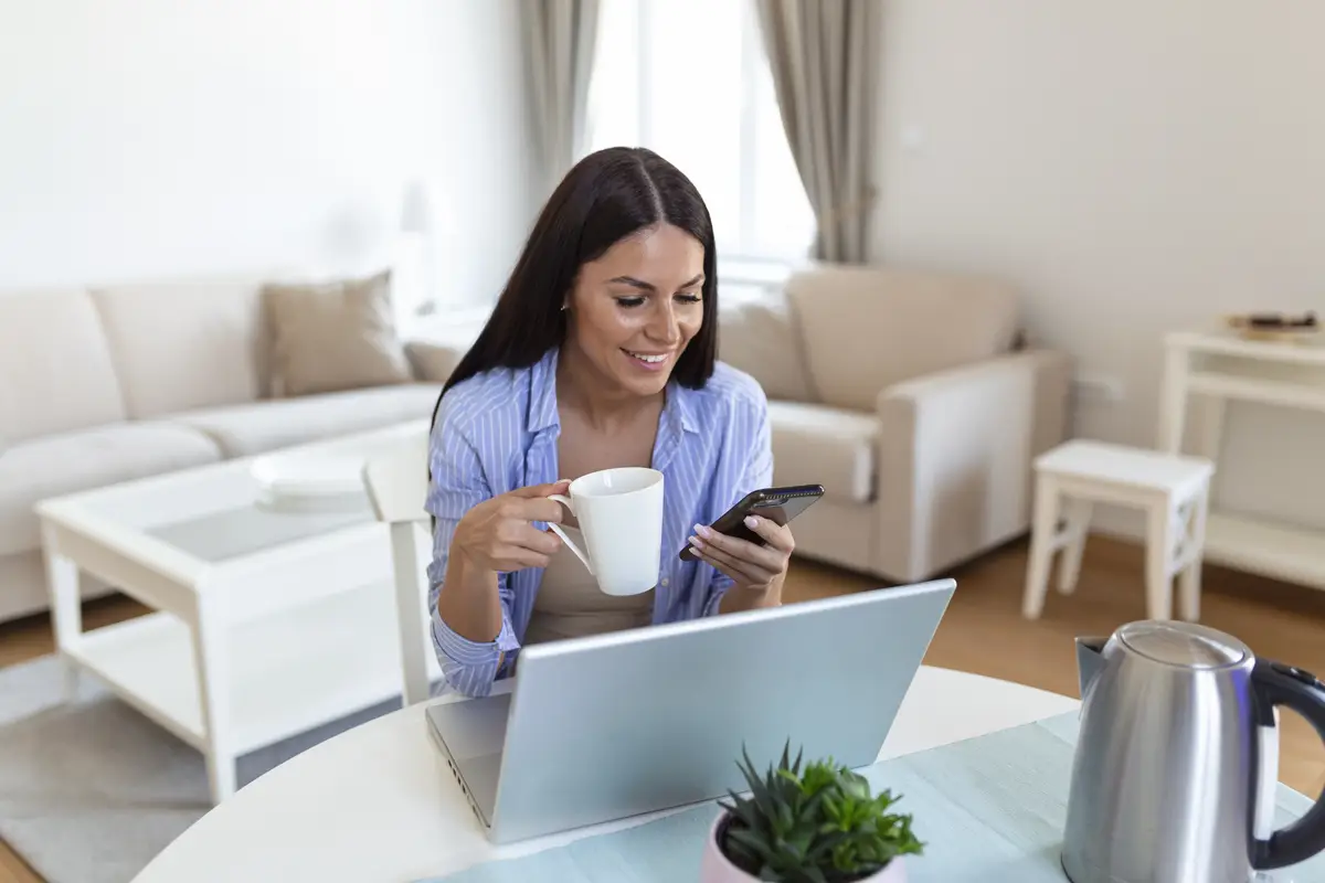 Self employed woman working with her phone and laptop at home with a cup of coffee Happy woman using mobile phone while working at home with laptop Smiling woman messaging with smartphone