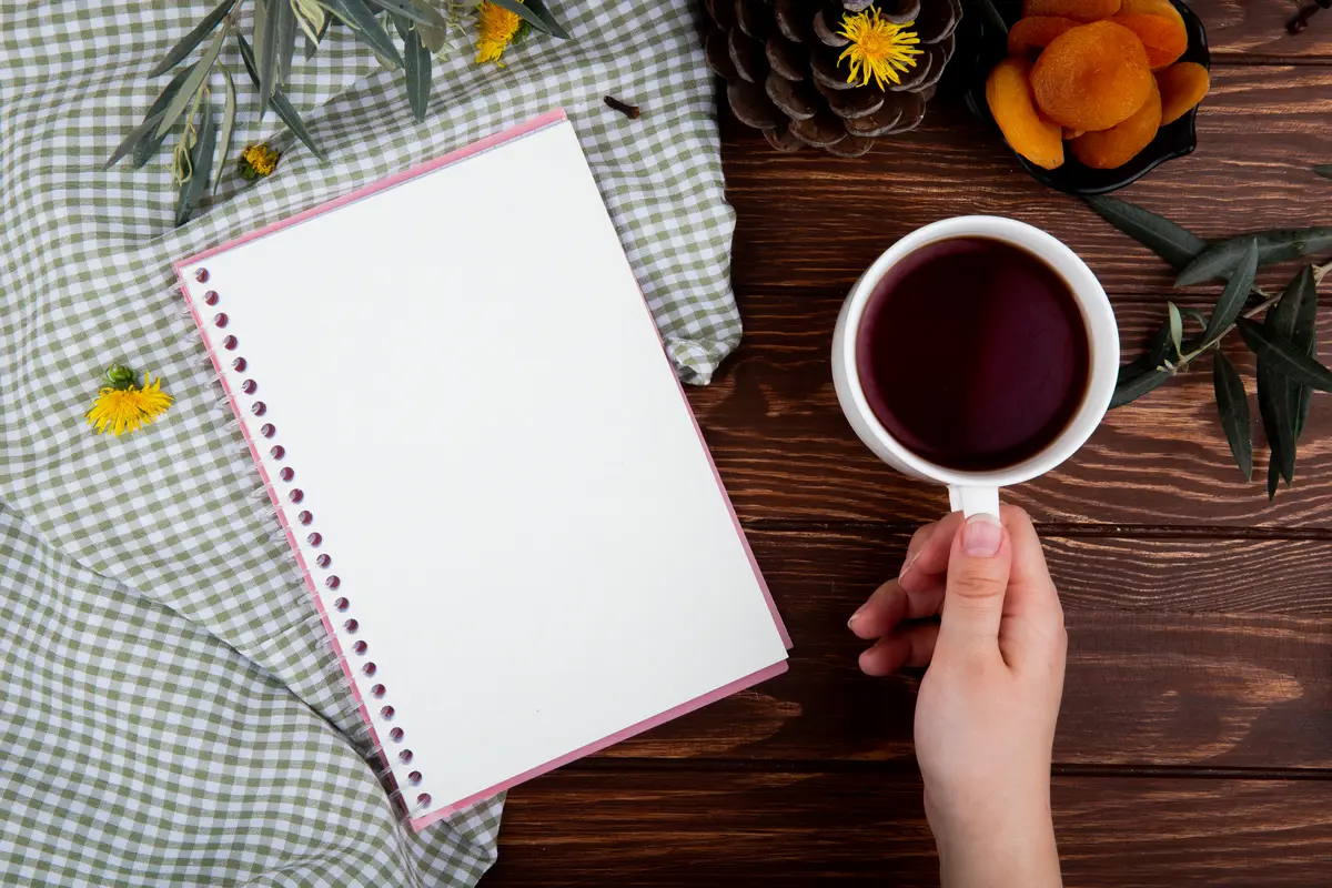 Top view of a hand holding a mug of tea with sketchbook on wood