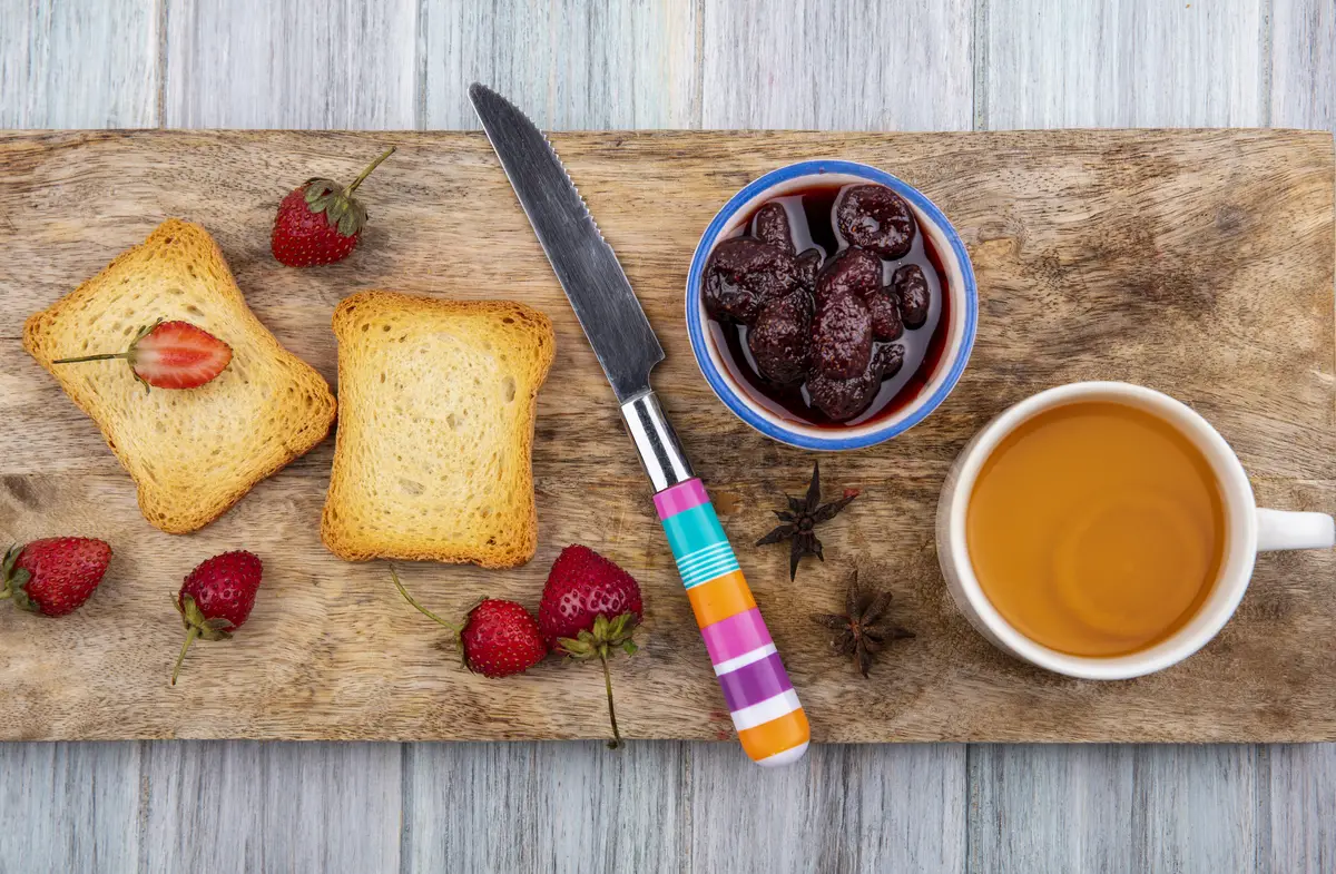 Top view of strawberry jam on a bowl on a wooden kitchen board with knife with toasted slices of bread with fresh strawberries on a grey wooden background