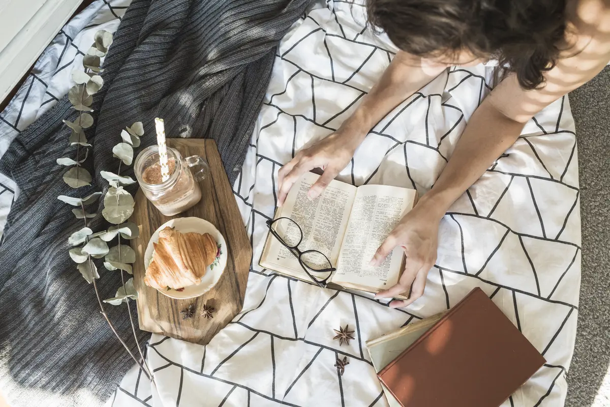 Anonymous woman reading book on bed near breakfast food