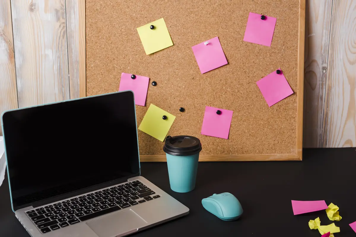 Laptop; takeaway coffee cup; mouse and corkboard with adhesive notes on black desk