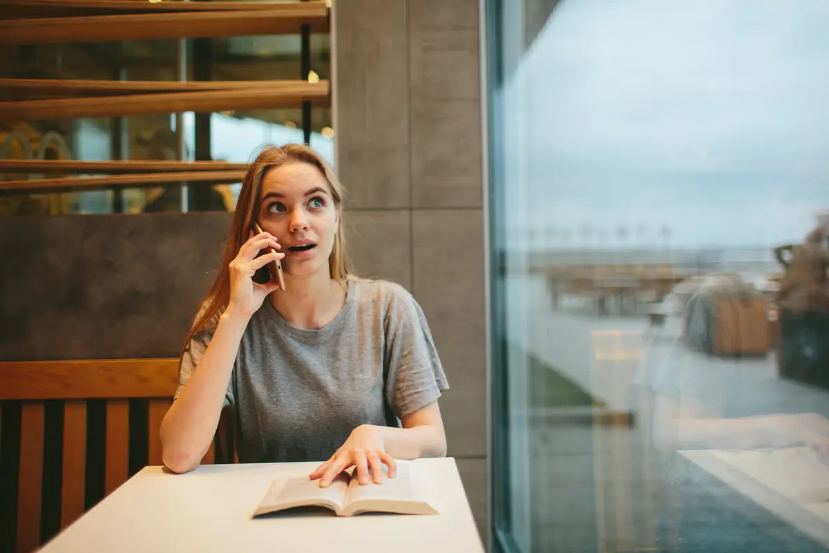 Blonde is reading a book and talking on the phone in a diner or restaurant