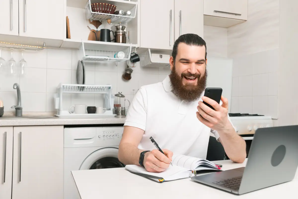Cheerful bearded man is video talking on the phone while writing in his planner.