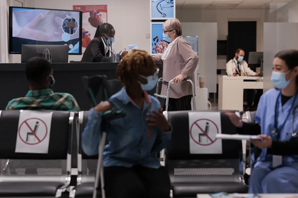 Mujer mayor con bastidor de marcha hablando con recepcionista en mostrador de recepción del hospital, esperando asistir a cita en instalación médica. Paciente con discapacidad física y mascarilla en área de espera.