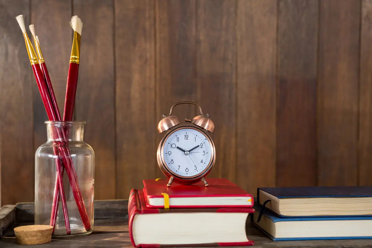 Books, alarm clock and paint brush arranged on wooden table
