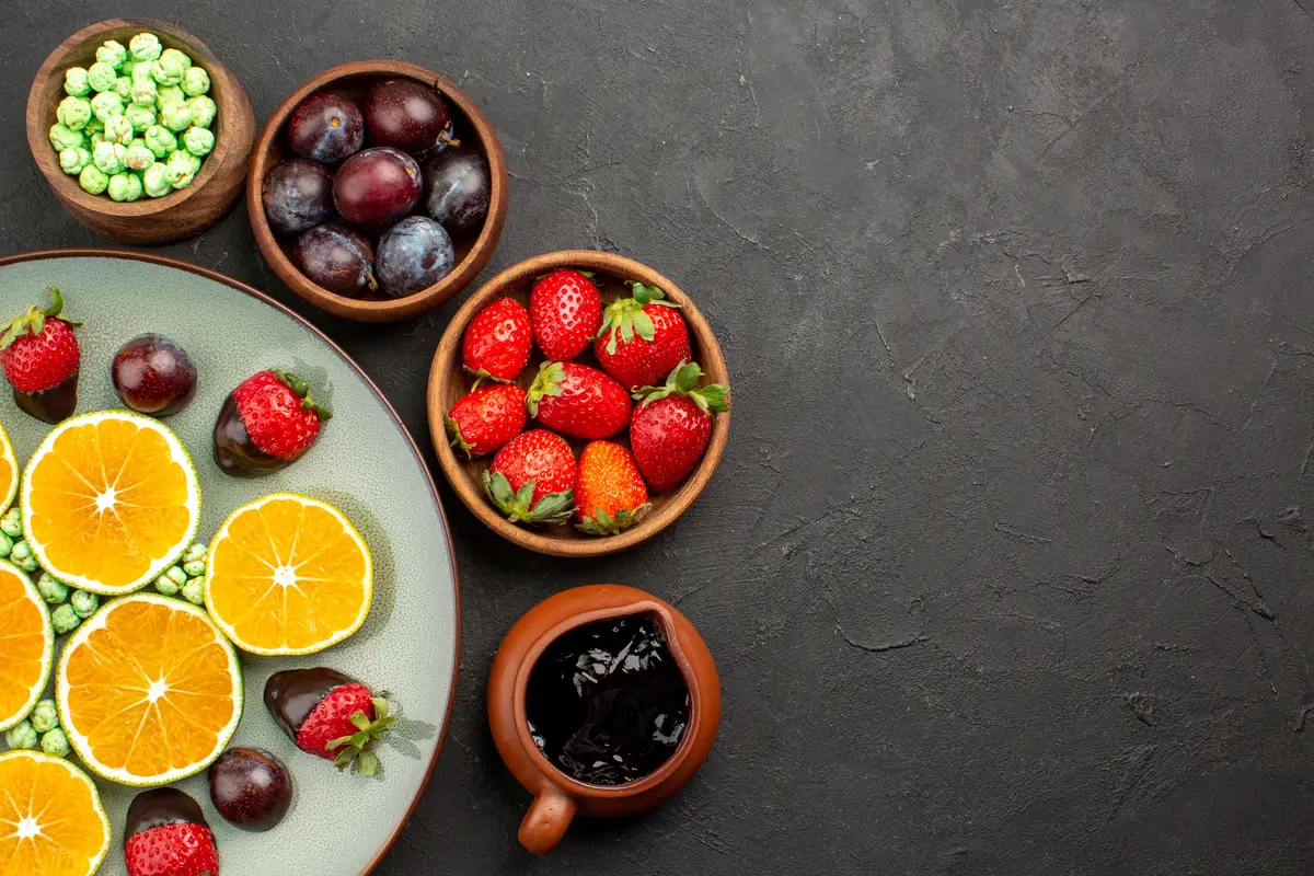 Top close-up view fruits green candies chocolate-covered strawberry chopped orange and bowls of berries and chocolate sauce on the table