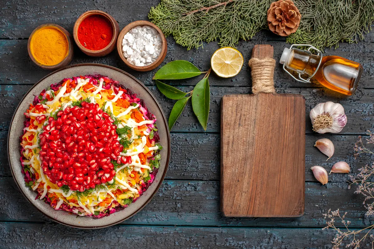 Top view Christmas dish dish with seeds of pomegranate bottle of oil next to the cutting board bowls of spices lemon garlic spruce branches with cones on the table