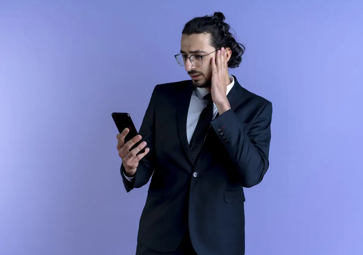 Business man in black suit and glasses looking at screen of his smartphone with confuse expression standing over blue wall