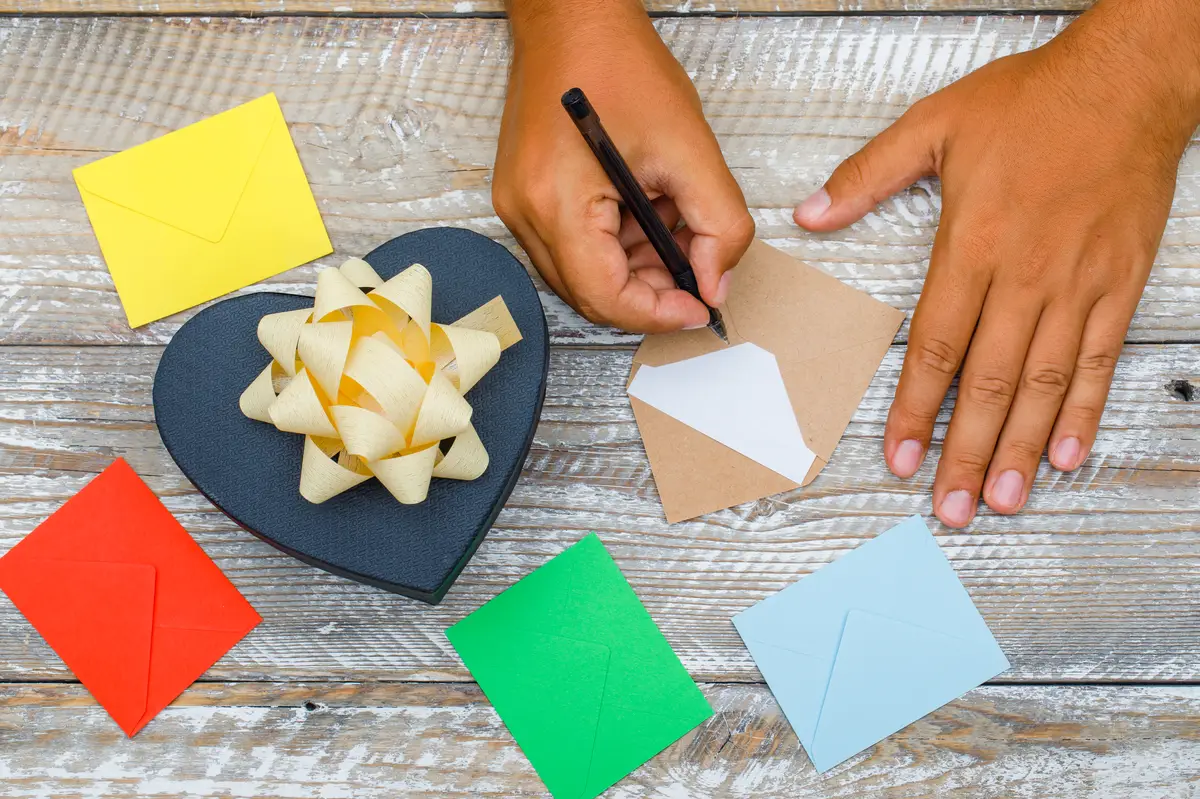 Birthday concept with gift box, envelopes on wooden background flat lay. man writing greeting card with pen.