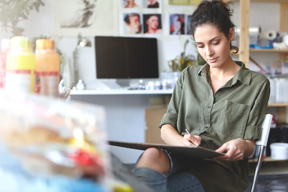 Female artist dressed casually, working on her sketch while drawing something and sitting in her workshop. Creative woman being involved in painting. People, hobby and creative process concept