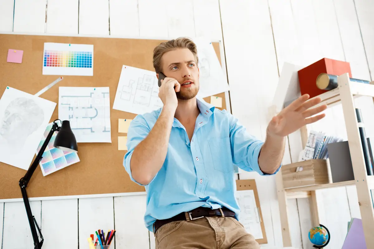 Young handsome confident businessman standing at table talking on phone. White modern office interior