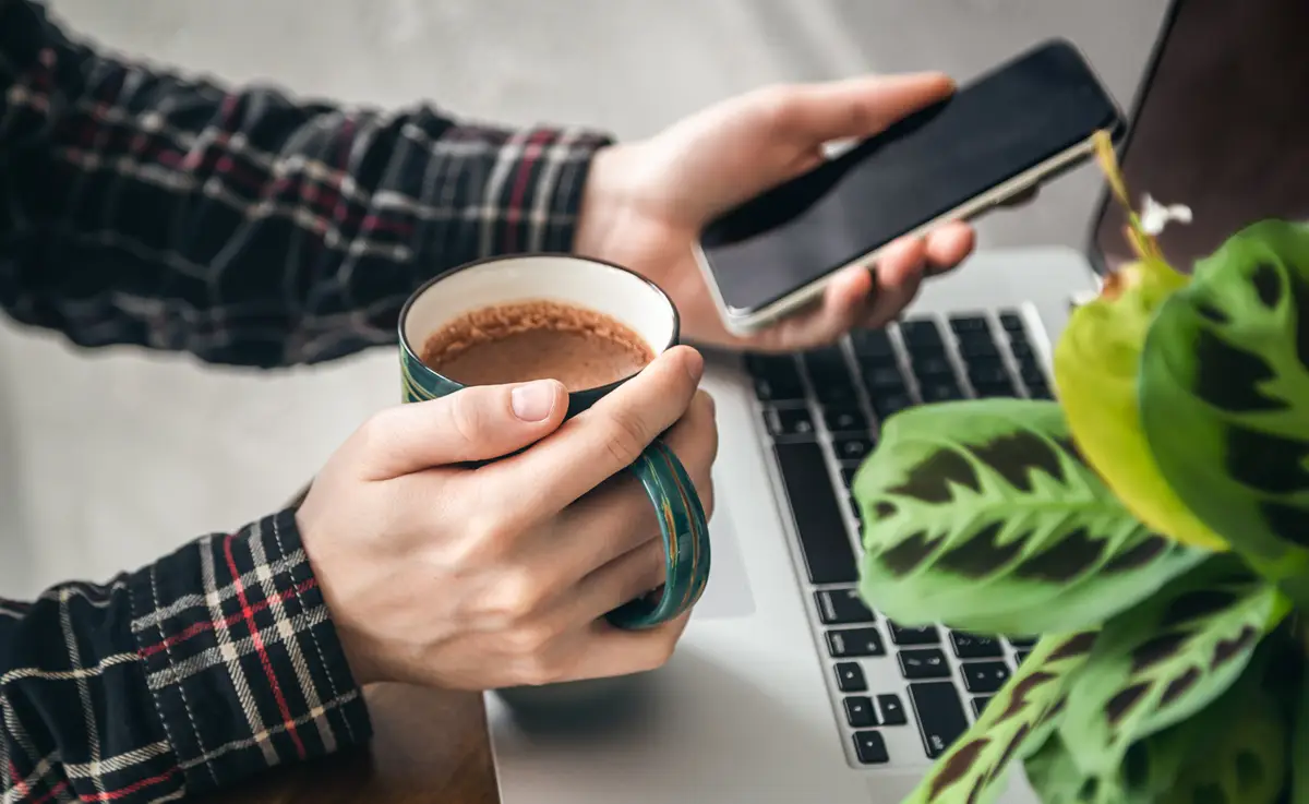 A cup of cocoa and a smartphone in the hands of a man in front of a laptop