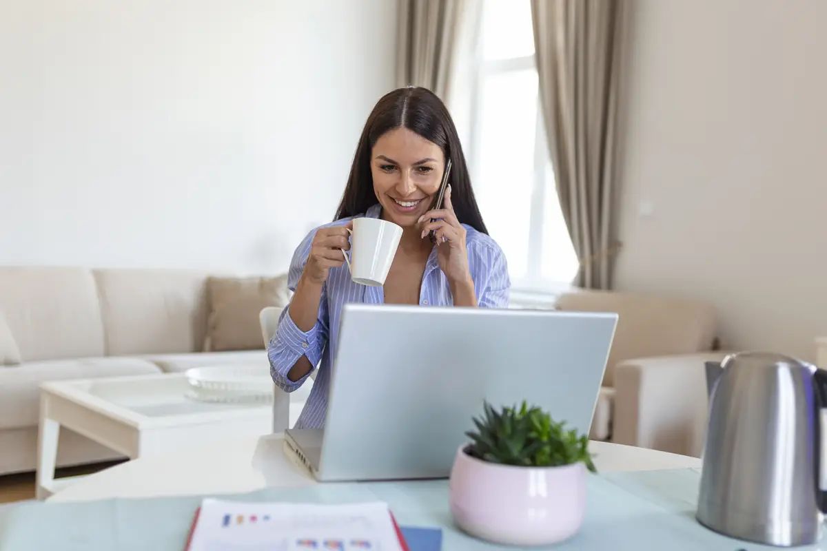 Banco Digital nos EUA para Brasileiros: Guia Completo de Abertura e Uso 2 Self employed woman working with her phone and laptop at home with a cup of coffee Happy woman using mobile phone while working at home with laptop Smiling woman messaging with smartphone