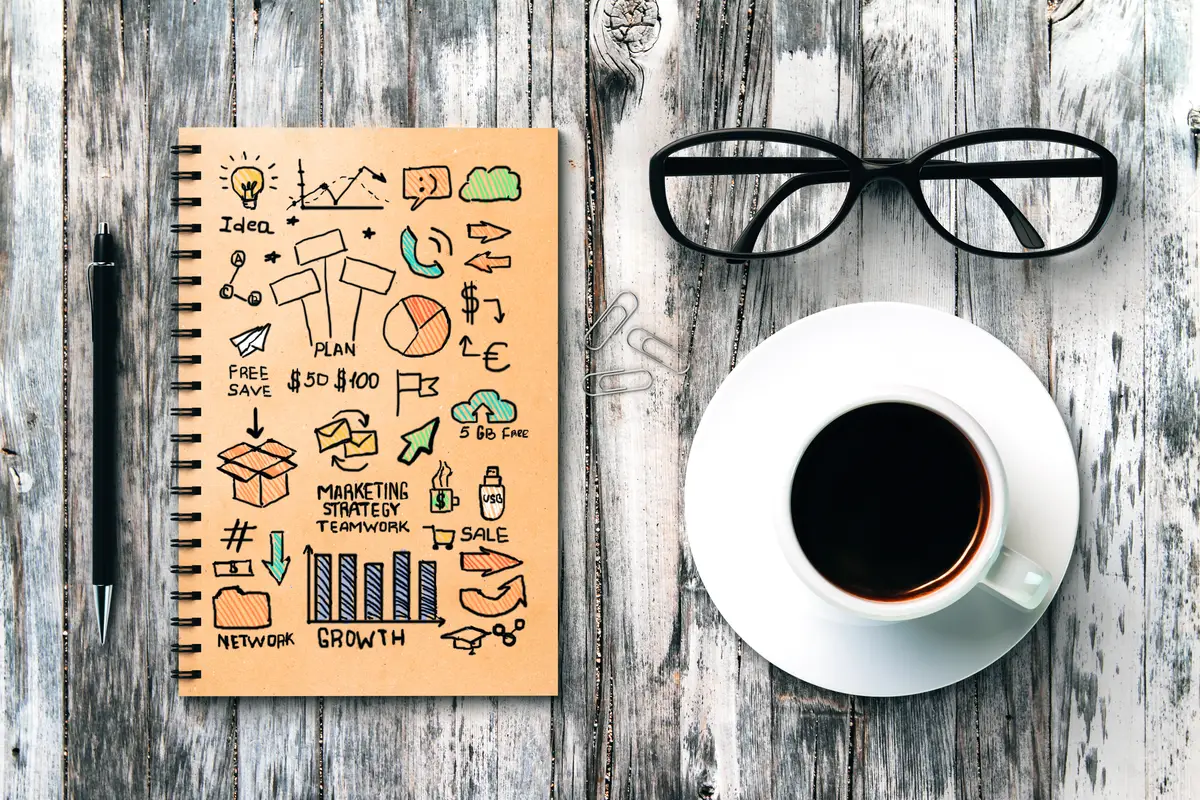 Top view of textured wooden desktop with business sketch in spiral notepad coffee cup glasses pen and paper clips