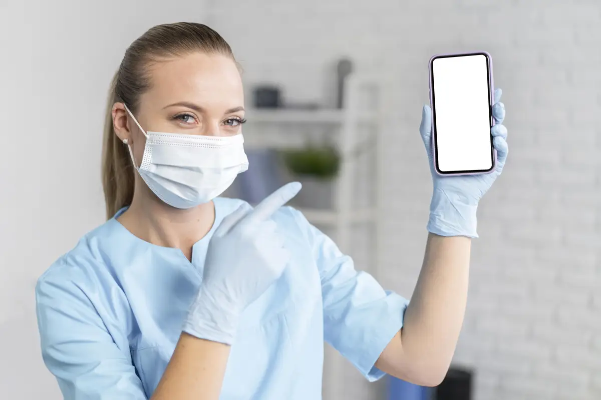 Female physiotherapist with medical mask holding and pointing at smartphone