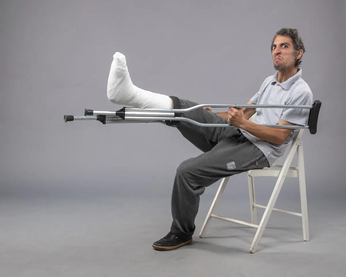 Front view young male sitting with broken foot using crutches for walking on a grey wall foot twist accident leg pain broken