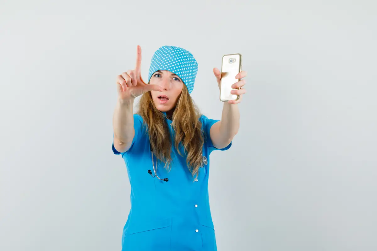 Female doctor holding mobile phone and gesturing with fingers in blue uniform 