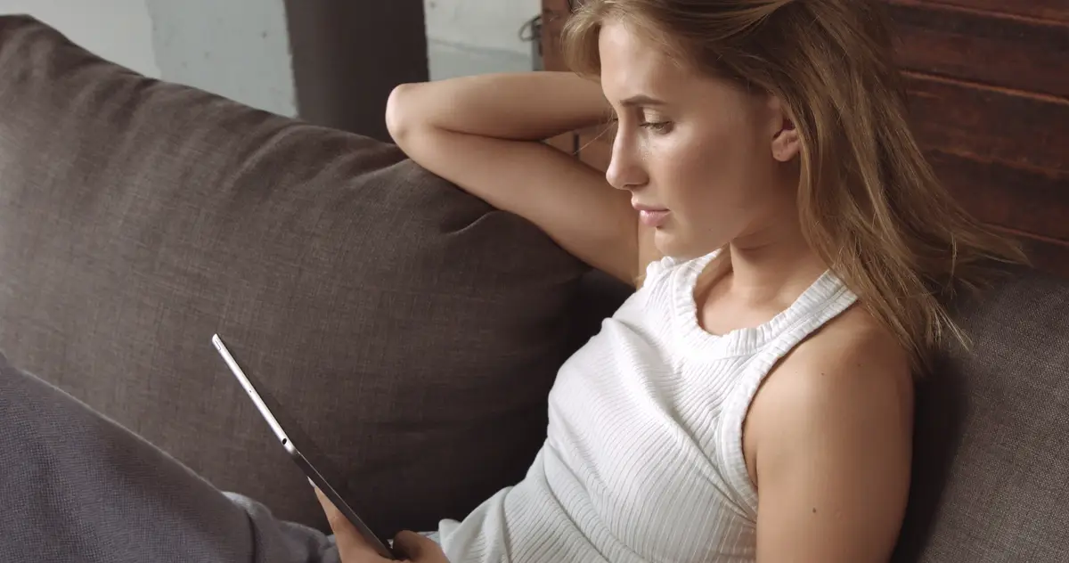 Hot blond model reading on her tablet while sitting on a dark gray couch covered with a soft throw