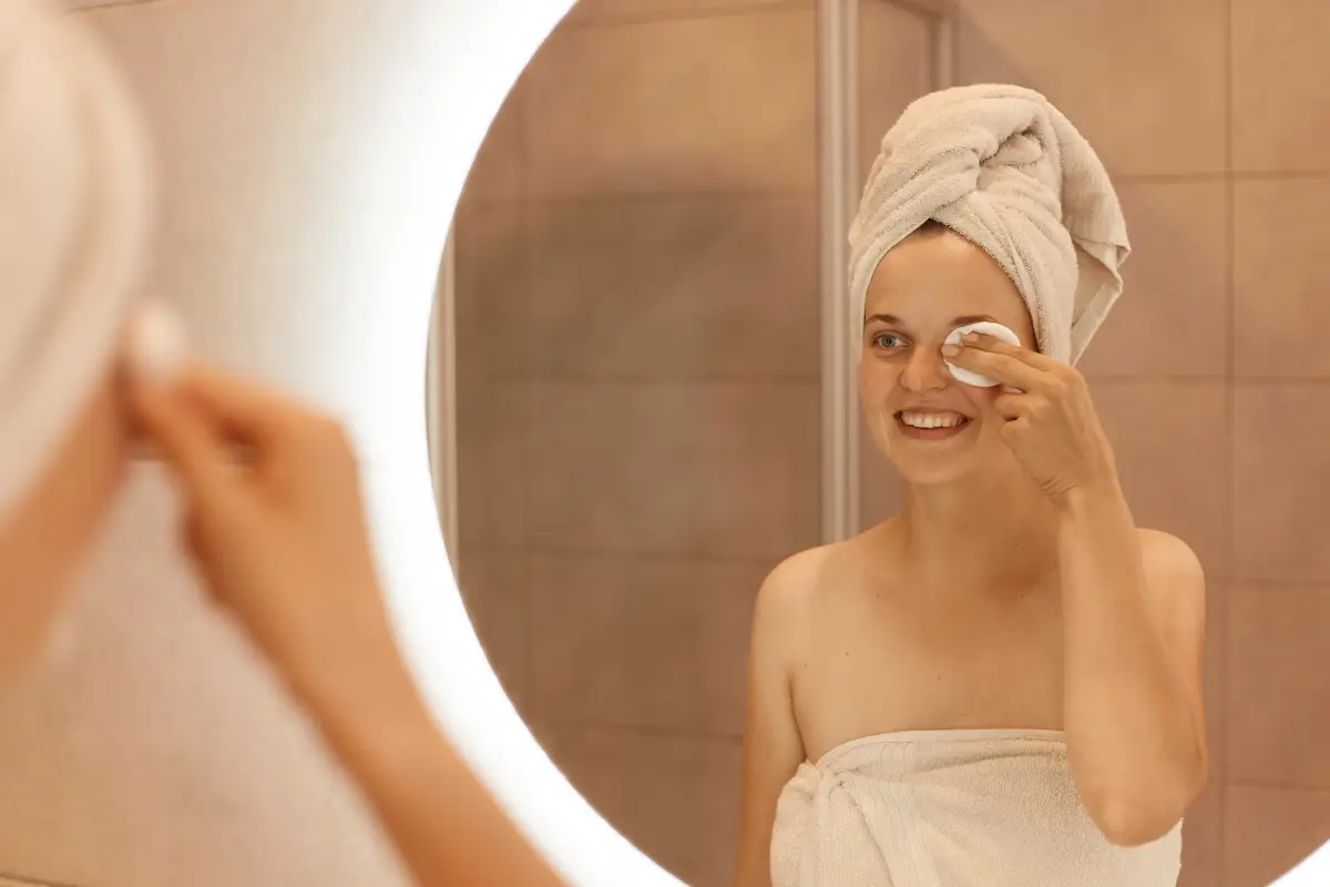 Portrait of beautiful Caucasian woman in towel on her head looking in mirror and cleaning her face with cotton pad, removing makeup from her eyes, smiling and expressing positive.