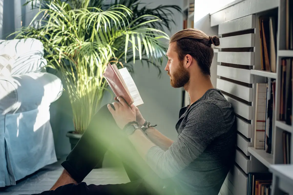 Stylish bearded male reading a book in a room with green plants.