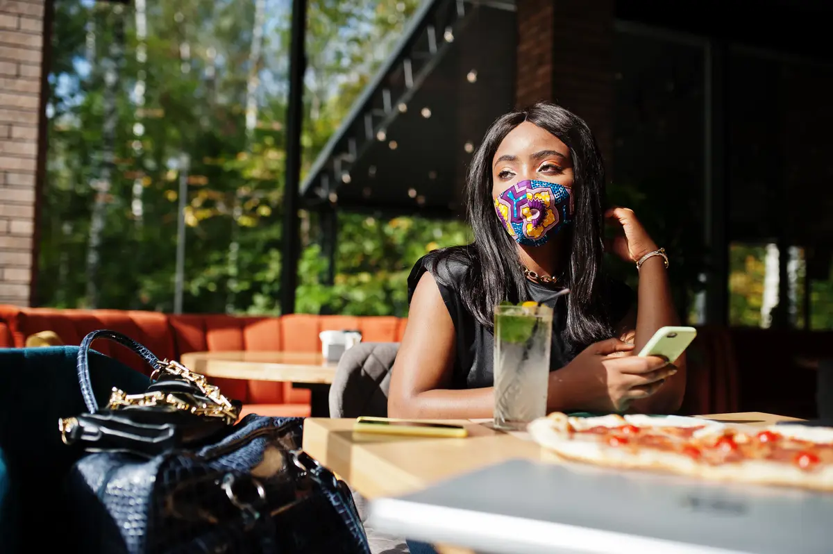 Fashionable african american woman wear hand made face mask sitting at restaurant look at mobile phone while eat pizza New normal life